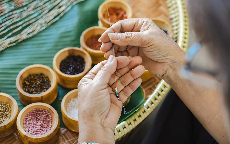 Close-up of hands threading a bead beside bowls of colored stones