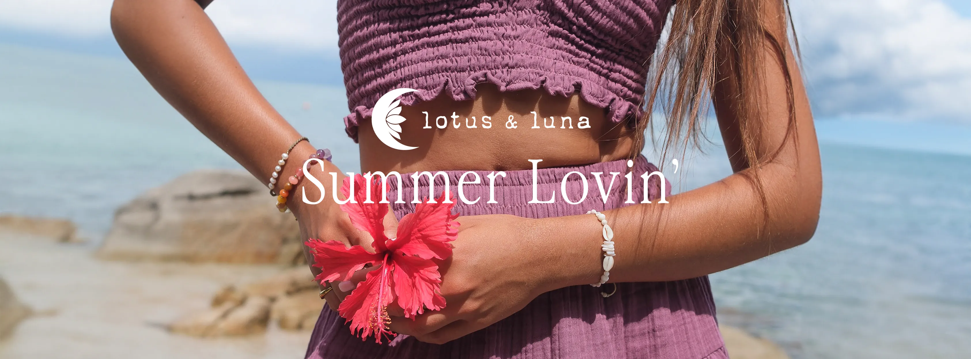 Lotus & Luna Summer Lovin' banner: a model in a purple set holding a red hibiscus at the shoreline